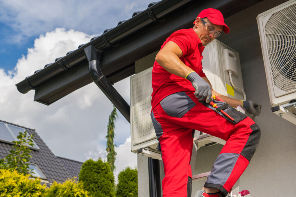 A technician in red and black uniform services an air conditioning unit.