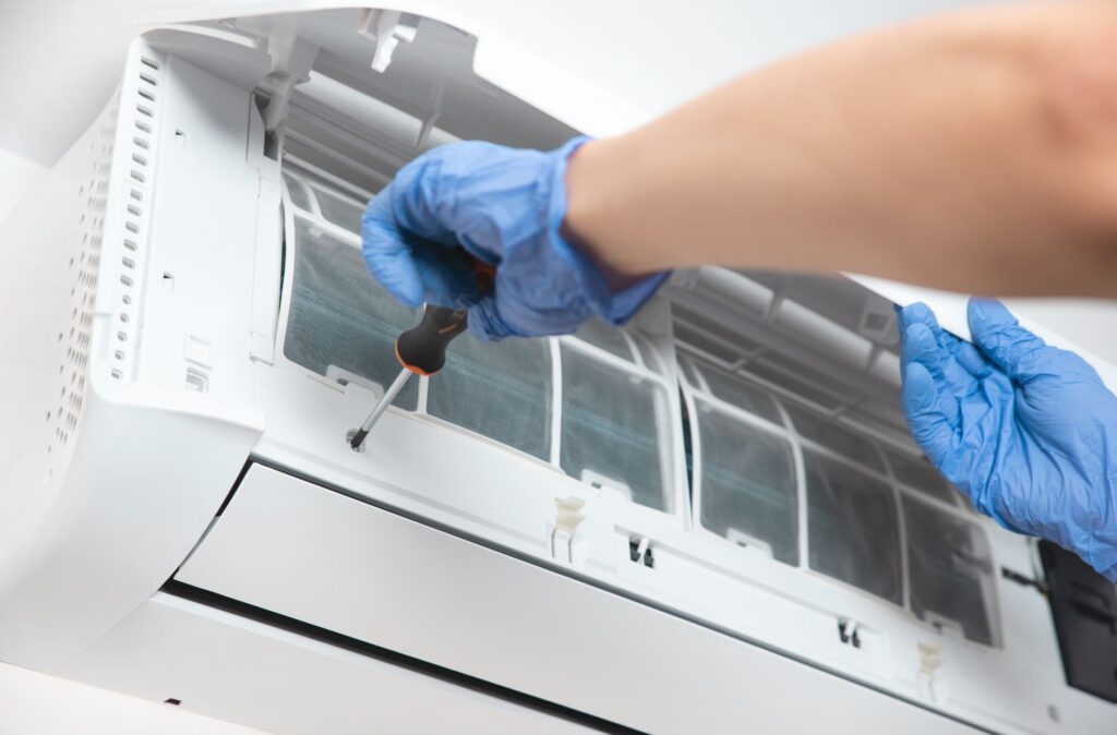 A person wearing blue gloves uses a screwdriver to repair or maintain a wall-mounted air conditioning unit.