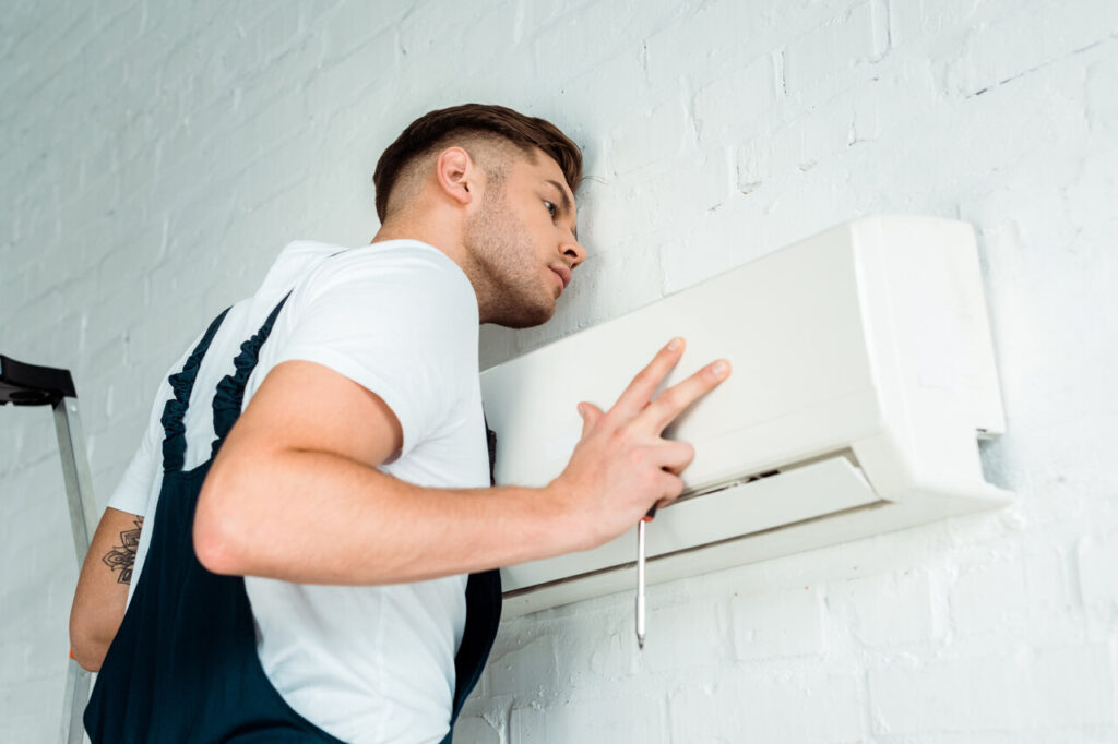 A technician examines a wall-mounted air conditioner closely, holding a tool.