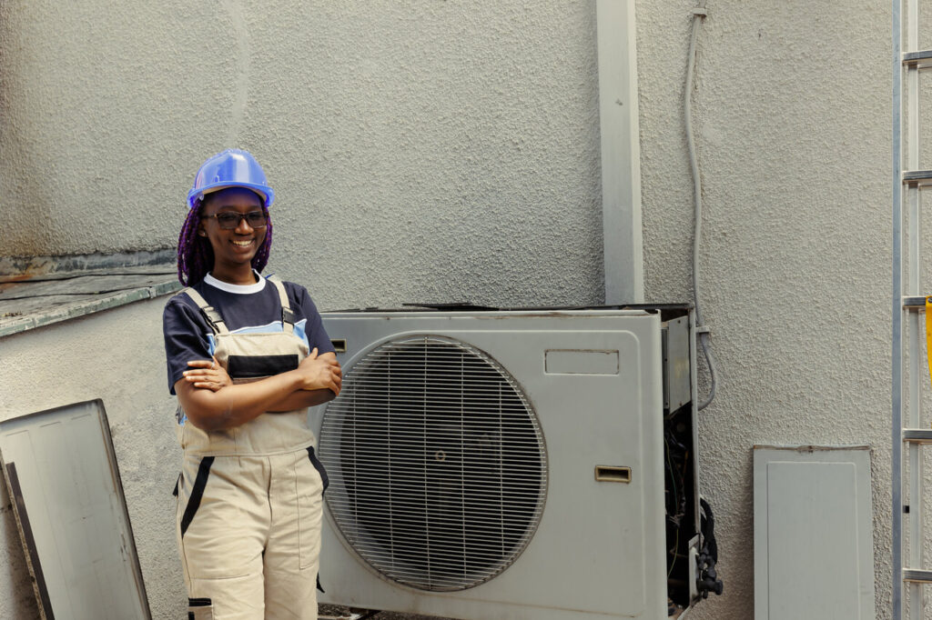 A woman wearing overalls and a hard hat stands next to an air conditioning unit.
