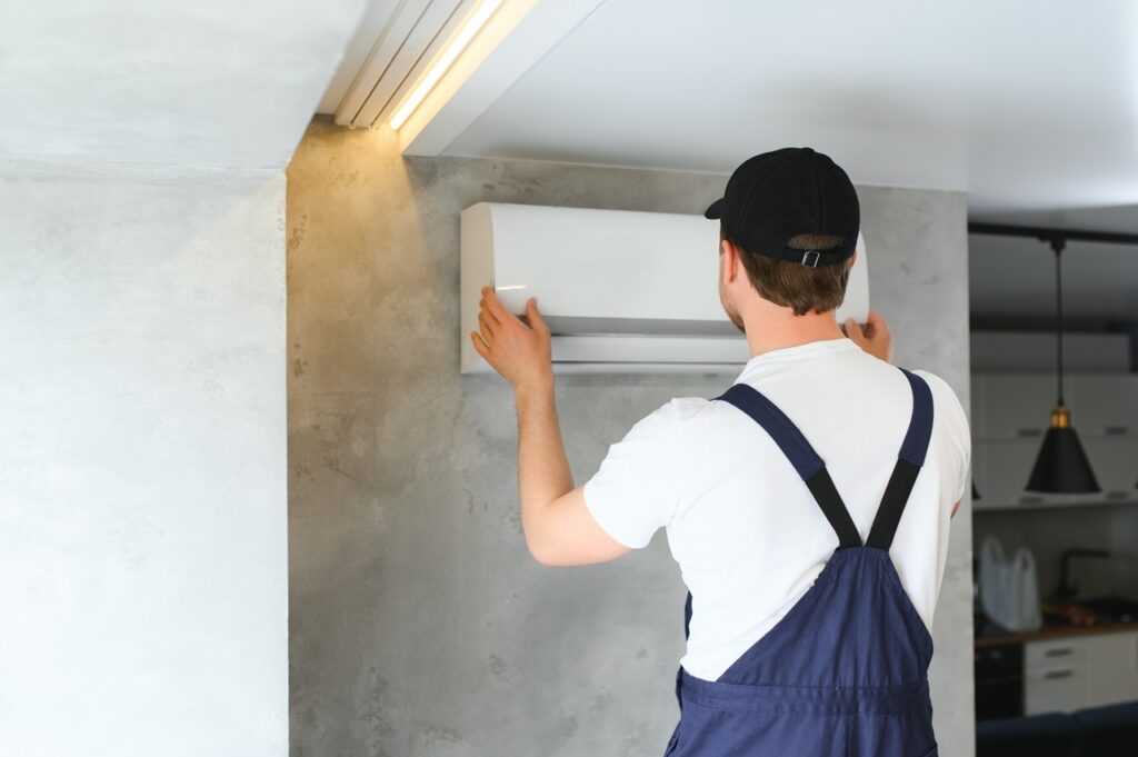 A man in overalls repairs an air conditioner, focused on the unit with tools in hand.