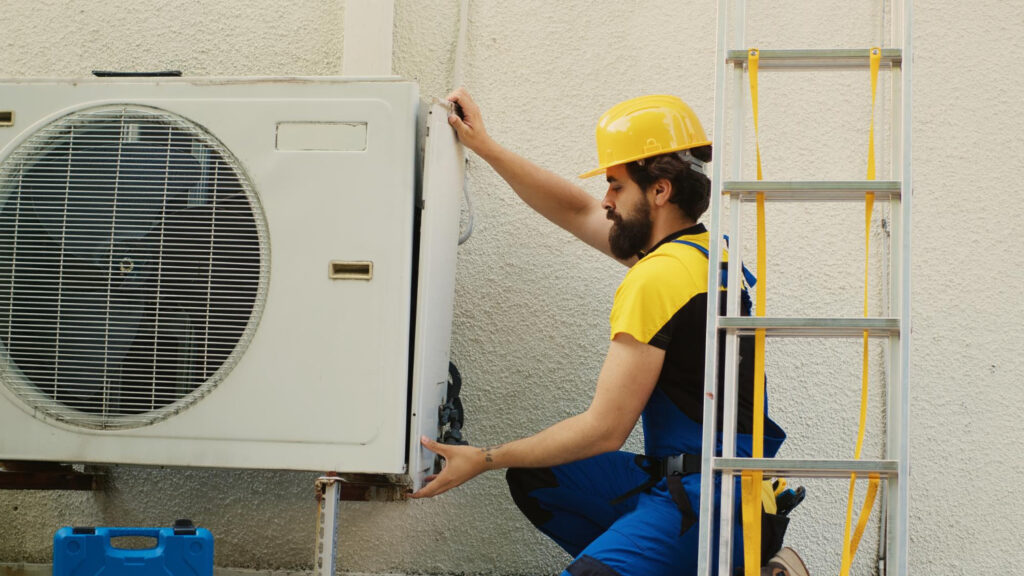 A man in blue overalls and a hard hat repairs an air conditioner.