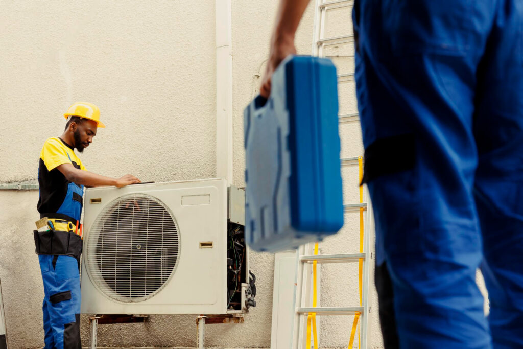 Two workers in blue overalls and hard hats are servicing an air conditioning unit.