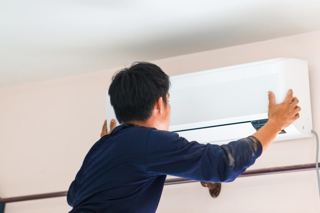 A technician in a blue shirt installs a white air conditioning unit on a light-colored wall.